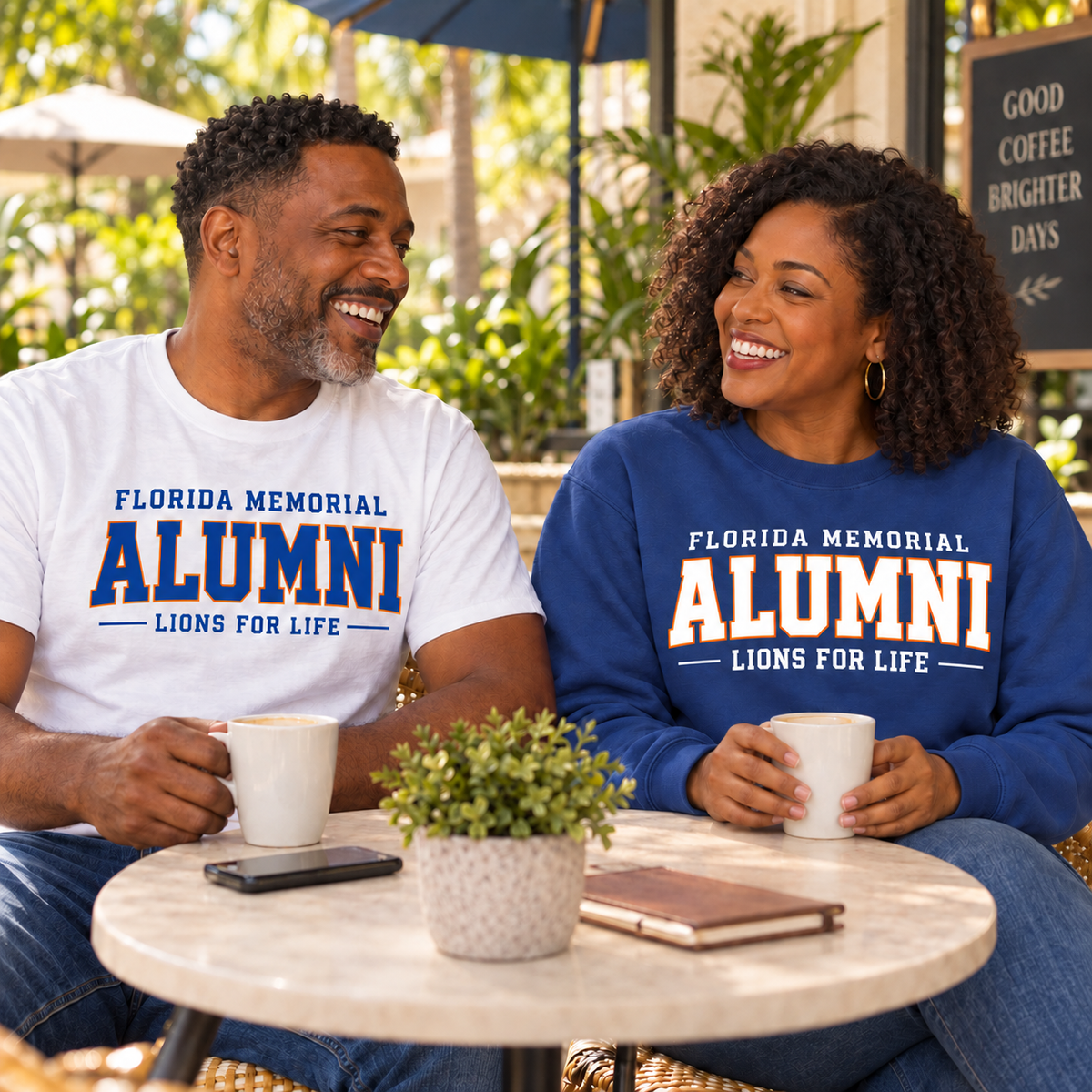 Florida Memorial University Lions for Life lifestyle mockup: two adults at an outdoor cafe wearing a white tee with blue-and-orange alumni lettering and a dark blue sweatshirt with white-and-orange lettering, both shown from the front.